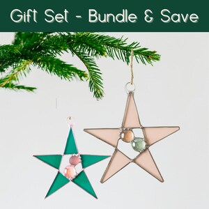 Puede incluir: Dos adornos en forma de estrella con cuentas de vidrio, uno verde y otro rosa, cuelgan de una cuerda. Los adornos están colocados sobre un fondo blanco con una ramita de hoja perenne. El texto "Gift Set - Bundle & Save" está en la parte superior.