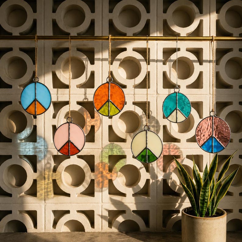 Six colorful stained glass peace sign suncatchers hanging from a brass rod against a sunlit mid-century breeze block wall, with soft reflections and a potted snake plant below.