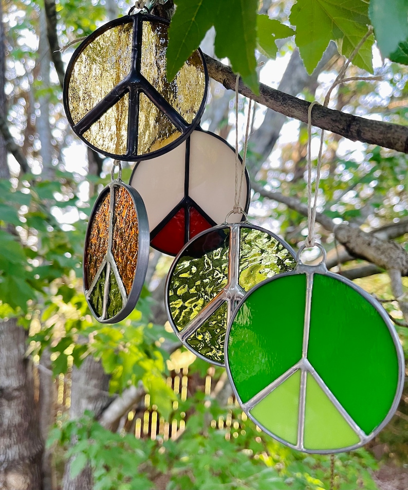 May include: Four stained glass peace sign ornaments hanging from a tree branch. The ornaments are in various colors, including gold, white, red, green, and brown.