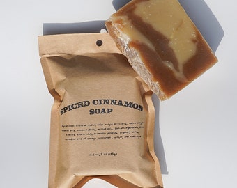 Op de afbeelding: Een bruine papieren zak met een wit label waarop "Spiced Cinnamon Soap" staat en de ingrediënten worden vermeld. De zak bevat een reep bruine en beige zeep met een swirl-patroon. De zeep weegt 142 g (5 oz).