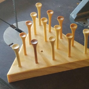 May include: A wooden golf tee holder in a triangular shape. The holder is made of light-colored wood and features several holes with golf tees inserted. The tees are a mix of light and medium brown colors.
