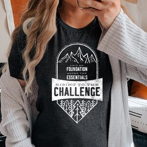 K&ouml;nnte beinhalten: Ein dunkelgraues T-Shirt mit einem wei&szlig;en Grafikdesign, das eine Bergkette und den Text "Laid my foundation, added the essentials, rising to the challenge" zeigt.