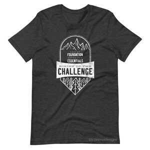 K&ouml;nnte beinhalten: Ein dunkel-melierter grauer T-Shirt mit einem wei&szlig;en Grafikdesign. Die Grafik zeigt eine Bergkette mit dem Text "Laid my foundation, added the essentials, rising to the challenge".