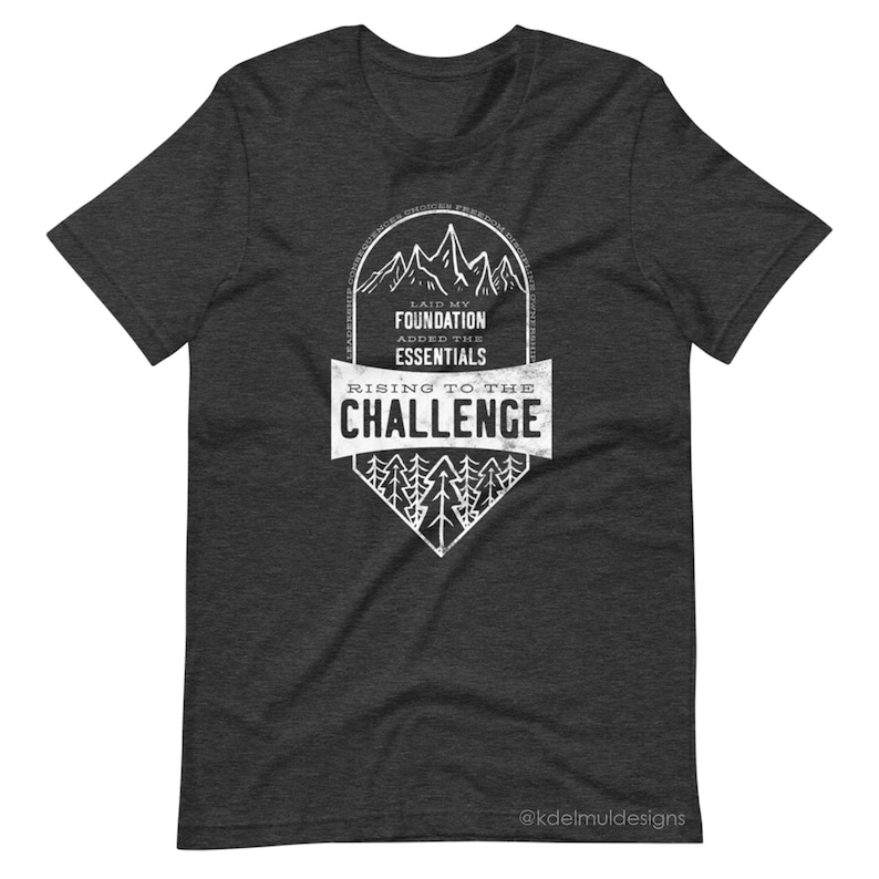 K&ouml;nnte beinhalten: Ein dunkel-melierter grauer T-Shirt mit einem wei&szlig;en Grafikdesign. Die Grafik zeigt eine Bergkette mit dem Text "Laid my foundation, added the essentials, rising to the challenge".