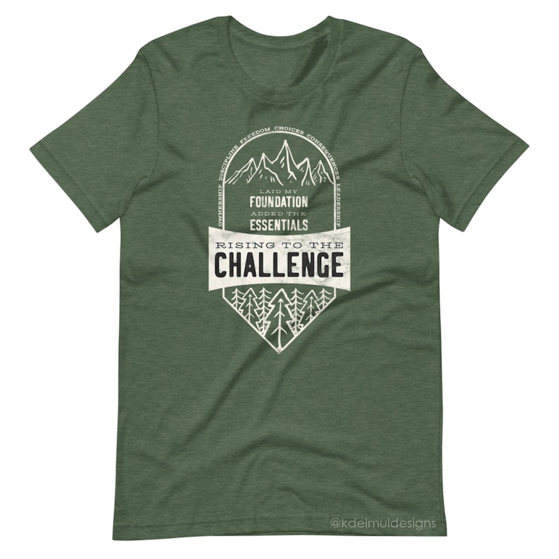 K&ouml;nnte beinhalten: Gr&uuml;nes T-Shirt mit einem wei&szlig;en Grafikdesign, das eine Bergkette und den Text "Laid my foundation, added the essentials, Rising to the challenge" zeigt. Die Grafik ist von einem Kreis mit dem Text "Discipline, Freedom, Choices, Consequences, Resilience" umgeben. Der Text "@kdelmuldesigns" befindet sich am unteren Rand des Shirts.