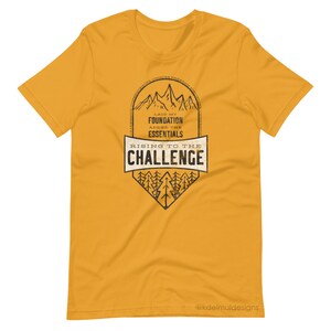 K&ouml;nnte beinhalten: Gelbes T-Shirt mit kurzen &Auml;rmeln und einem schwarz-wei&szlig;en Grafikdesign. Die Grafik zeigt eine Bergkette mit dem Text "Laid my foundation, added the essentials, rising to the challenge". Die Grafik enth&auml;lt auch eine Waldszene mit B&auml;umen. Der Text "@kdelmuldesigns" befindet sich am unteren Rand der Grafik.