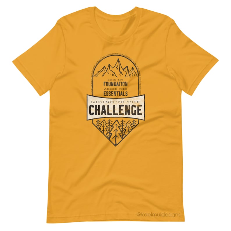 K&ouml;nnte beinhalten: Gelbes T-Shirt mit kurzen &Auml;rmeln und einem schwarz-wei&szlig;en Grafikdesign. Die Grafik zeigt eine Bergkette mit dem Text "Laid my foundation, added the essentials, rising to the challenge". Die Grafik enth&auml;lt auch eine Waldszene mit B&auml;umen. Der Text "@kdelmuldesigns" befindet sich am unteren Rand der Grafik.