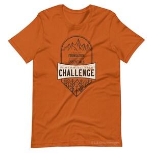 K&ouml;nnte beinhalten: Orangefarbenes T-Shirt mit einem schwarzen Grafikdesign einer Bergkette mit B&auml;umen und dem Text "Laid my foundation, added the essentials, Rising to the challenge".