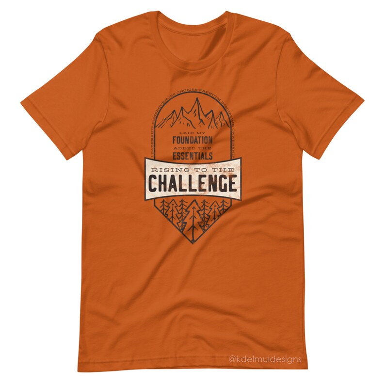K&ouml;nnte beinhalten: Orangefarbenes T-Shirt mit einem schwarzen Grafikdesign einer Bergkette mit B&auml;umen und dem Text "Laid my foundation, added the essentials, Rising to the challenge".