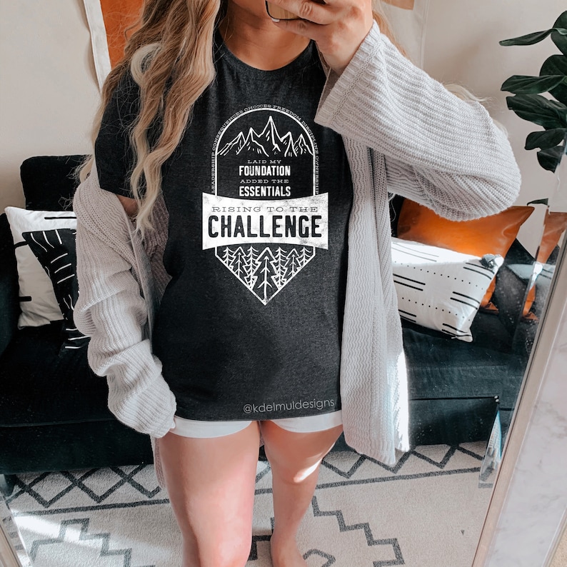 K&ouml;nnte beinhalten: Ein dunkelgraues T-Shirt mit einem wei&szlig;en Grafikdesign, das eine Bergkette und den Text "Laid my foundation, added the essentials, rising to the challenge" zeigt.