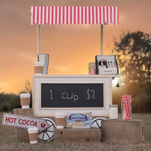 May include: A red and white striped awning over a wooden hot cocoa stand with a chalkboard sign that says "1 cup $2". There are cups of hot cocoa, a cash register, and a box of Swiss Miss marshmallows on the stand. The stand is on a wooden platform in front of a grassy field.