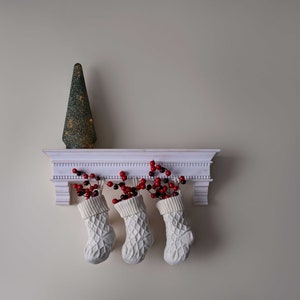 May include: Three white stockings with red berry sprigs hanging from a white shelf with a small green Christmas tree on top.