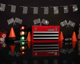 Op de afbeelding: Een rode gereedschapskist met drie laden en het woord "CRAFTSMAN" op de voorkant. De gereedschapskist is omringd door zwart-wit geblokte vlaggen, verkeerskegels, een benzinepomp en een blikje motorolie. De scène wordt weerspiegeld op een zwart oppervlak.