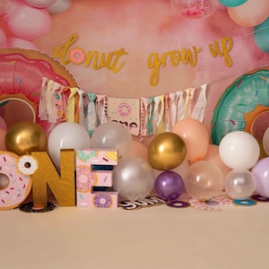 Puede incluir: Un telón de fondo de fiesta de primer cumpleaños con temática de donas con una pancarta que dice "donut grow up". El telón de fondo presenta un anillo inflable en forma de dona rosa y verde, un gran letrero "ONE" dorado y una variedad de globos coloridos.