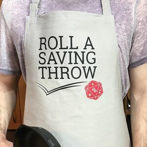 May include: A gray apron with the text "Roll a Saving Throw" and a red 20-sided die graphic.