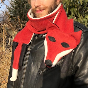 May include: A red fleece scarf with a white trim and a fox face design. The fox has black eyes and a black nose.