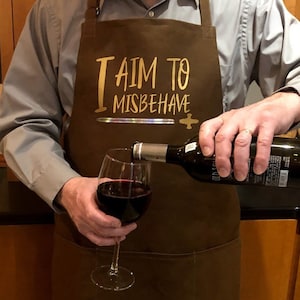 May include: Brown apron with the text "I AIM TO MISBEHAVE" in gold lettering. A person is pouring red wine into a wine glass. The apron has a sword and cross design.