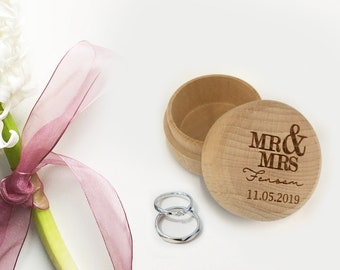May include: A wooden ring box with the text "MR & MRS Jensen 11.05.2019" engraved on the lid. Two silver wedding bands are next to the box.