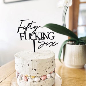 May include: A white cake with a layer of colorful candy eggs and a black cake topper that says "Fifty Fucking Six".