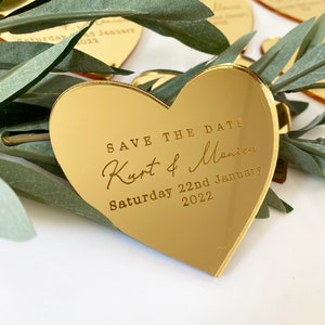 Puede incluir: Espejo dorado en forma de corazón con el texto "Save the Date" y "Kurt & Monica" en letra cursiva. La fecha "Saturday 22nd January 2022" está impresa debajo de los nombres.