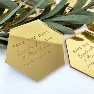 May include: Gold hexagonal mirror save the date cards with the text "Save the Date" and the names "Lesley Marie & Matthew" and the date "10th April 2021".