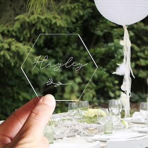 May include: A clear acrylic hexagon place card with the name "Hayley" written in cursive script. The place card is held in a hand against a backdrop of a white table with white chairs and a white paper lantern.