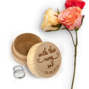 Könnte beinhalten: Eine hölzerne Ringbox mit der Aufschrift "with this ring A♥F 26.09.2019". Die Box ist geöffnet und zwei silberne Ringe befinden sich darin.