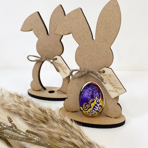 May include: Two wooden bunny-shaped Easter egg holders with a Cadbury Creme Egg in one. The holders have a tag with the names "Allie" and "Emily" written on them.
