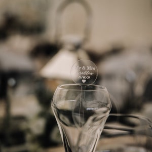 May include: A clear glass champagne flute with a round, clear acrylic wedding topper that says "Mr. & Mrs. [Name] 01/01/18" with a small heart.