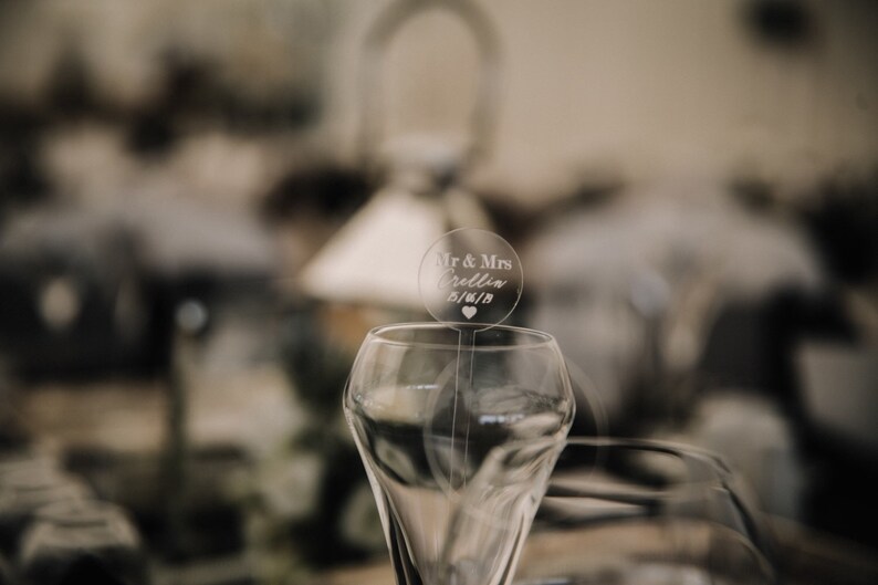 May include: A clear glass champagne flute with a round, clear acrylic wedding topper that says "Mr. & Mrs. [Name] 01/01/18" with a small heart.
