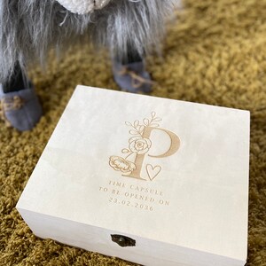 May include: A white wooden box with a floral design and the letter "P" engraved on the lid. The box has a brass latch and the text "Time Capsule to be opened on 23.02.2036" is engraved on the lid.