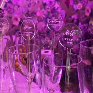May include: Clear acrylic place cards with names and the text "Liverpool 2019" on each card. The cards are placed in champagne flutes.