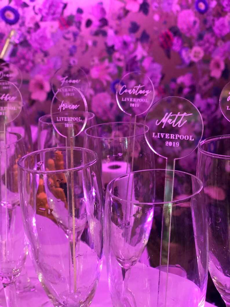 May include: Clear acrylic place cards with names and the text "Liverpool 2019" on each card. The cards are placed in champagne flutes.