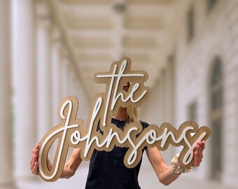 May include: A wooden sign with the text "The Johnsons" in white cursive lettering. The sign is held in front of a person's face.