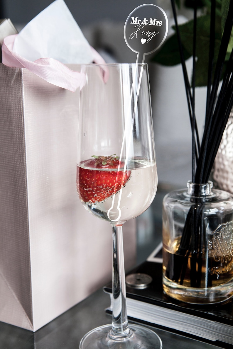 Clear perspex cocktail stirrer with personalized naming in a champagne flute with a strawberry. All with a gift bag in the background
