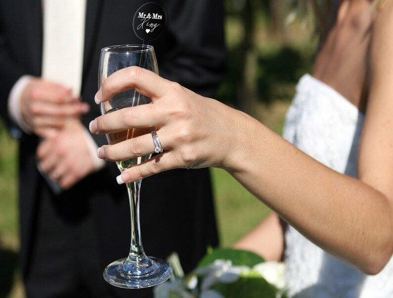 May include: A bride holds a champagne flute with a wedding topper that reads "Mr. & Mrs. King" with a heart symbol. The bride is wearing a white strapless wedding dress and a diamond ring on her finger.