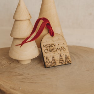 May include: Wooden Christmas bauble with a red ribbon. The bauble is shaped like an arch and has the text "Merry Christmas" and a design of three stylised Christmas trees.