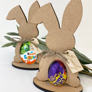 May include: Two wooden bunny rabbit cutouts with a chocolate egg in each. The bunny on the left has a Kinder Surprise egg and the bunny on the right has a Cadbury Creme Egg. Each bunny has a tag with a name on it, "Alice" and "Emily".