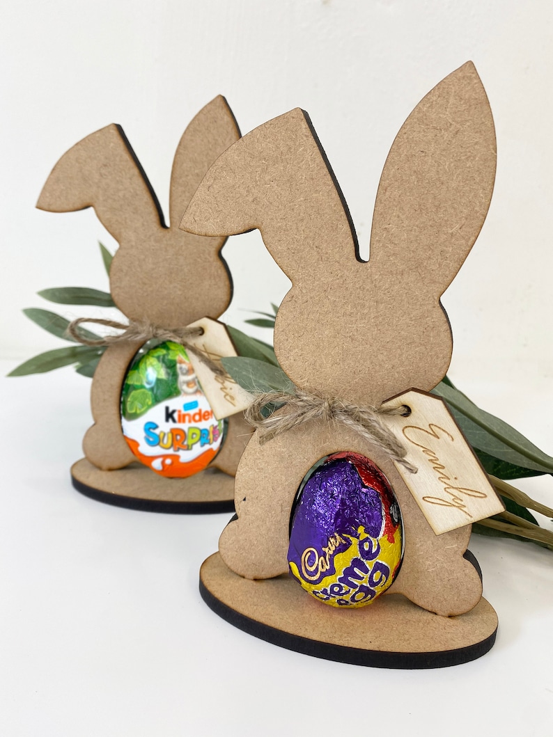 May include: Two wooden bunny rabbit cutouts with a chocolate egg in each. The bunny on the left has a Kinder Surprise egg and the bunny on the right has a Cadbury Creme Egg. Each bunny has a tag with a name on it, "Alice" and "Emily".
