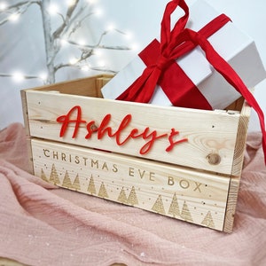 May include: A wooden Christmas Eve box with the name "Ashley's" in red script. The box contains a white gift with a red ribbon. The box has engraved Christmas tree designs and the words "CHRISTMAS EVE BOX".