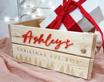May include: A wooden Christmas Eve box with the name "Ashley's" in red script. The box contains a white gift with a red ribbon. The box has engraved Christmas tree designs and the words "CHRISTMAS EVE BOX".