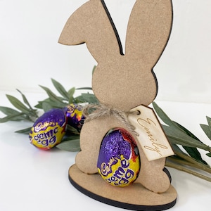May include: Wooden bunny rabbit cutout with a chocolate egg inside. The bunny has a tag that says "Emily".