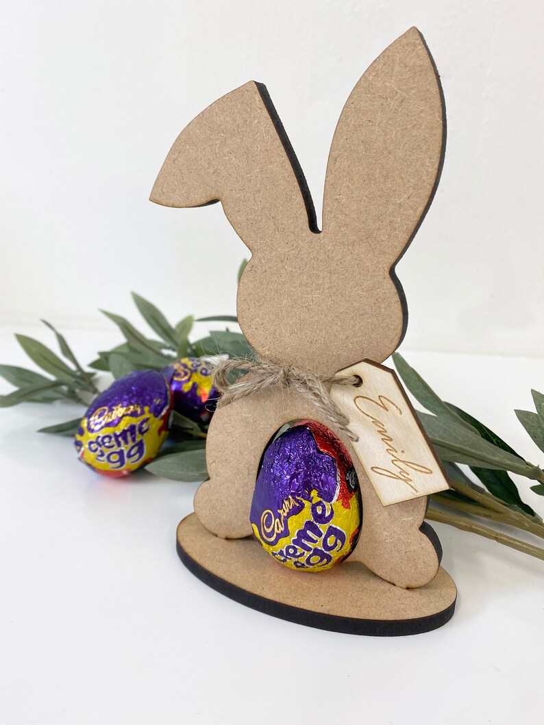 May include: Wooden bunny rabbit cutout with a chocolate egg inside. The bunny has a tag that says "Emily".