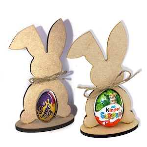 May include: Two wooden bunny rabbit cutouts with a small chocolate egg in the center of each. The bunny on the left has a purple and gold foil wrapped egg with the text "Cadbury Creme Egg". The bunny on the right has a green, orange, and white foil wrapped egg with the text "Kinder Surprise".