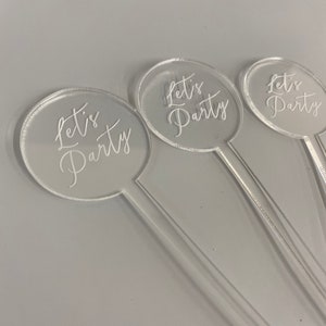 May include: Three clear acrylic drink stirrers with a round, clear acrylic topper that says "Let's Party" in white script.