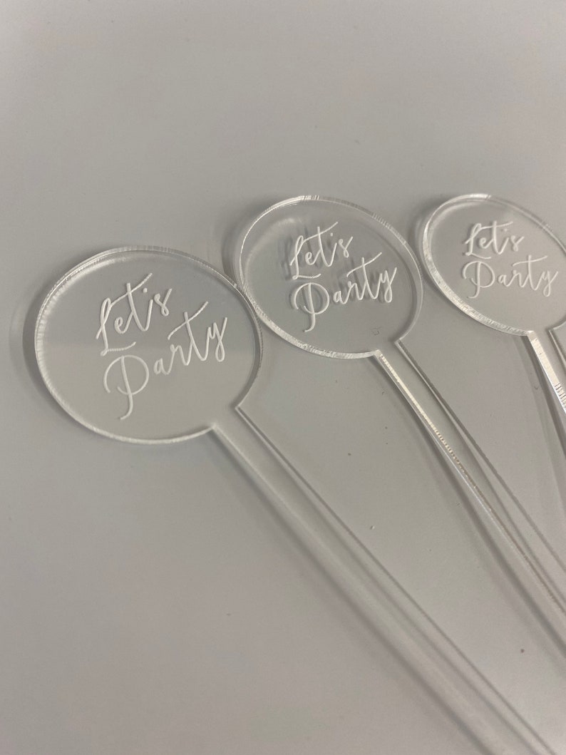 May include: Three clear acrylic drink stirrers with a round, clear acrylic topper that says "Let's Party" in white script.