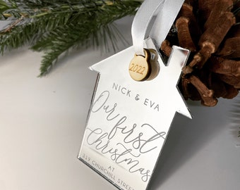 May include: A house-shaped mirror acrylic ornament featuring a white ribbon, a small wooden charm engraved with 2022, and printed text reading NICK & EVA, Our first Christmas, and 319 CHURCHILL STREET W. The ornament has a reflective surface.