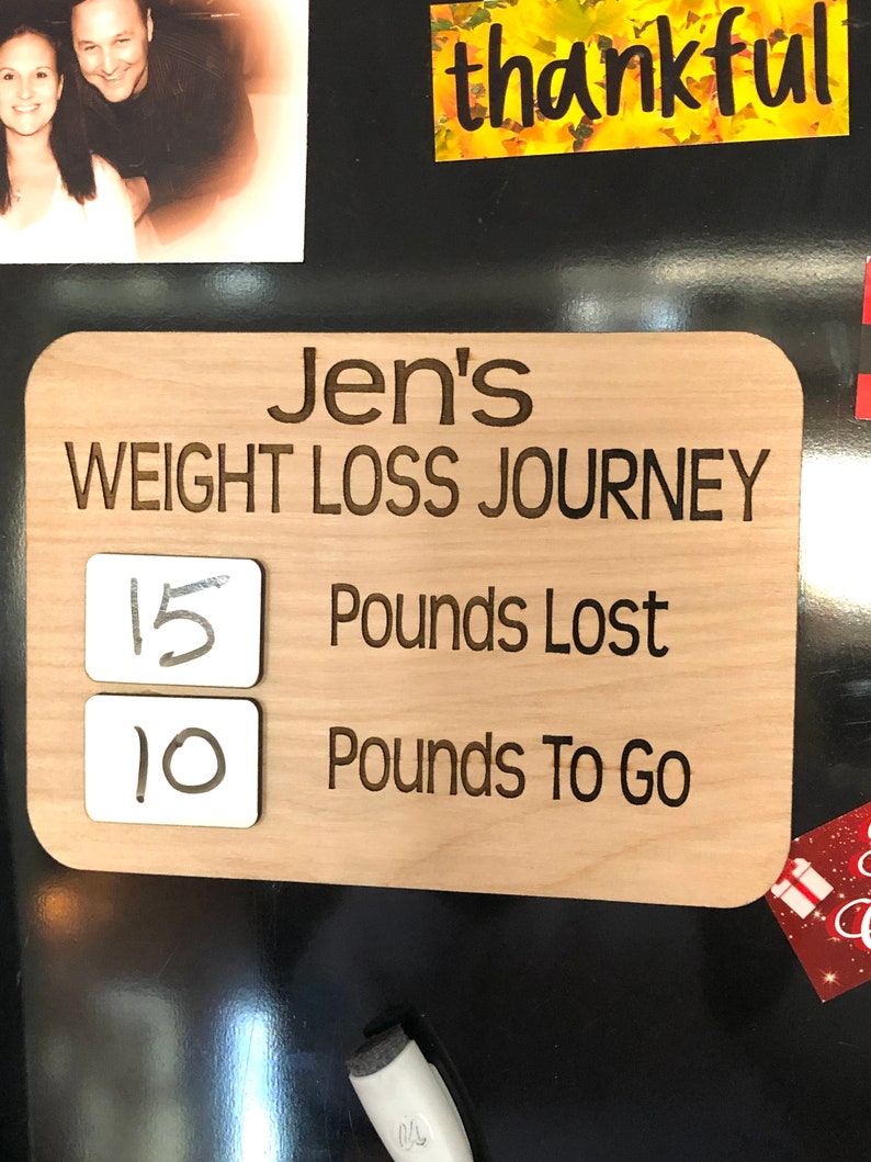 May include: A wooden sign with the text "Jen's Weight Loss Journey" and two white squares with the numbers "15" and "10" written on them. The text "Pounds Lost" and "Pounds To Go" are written below the squares.