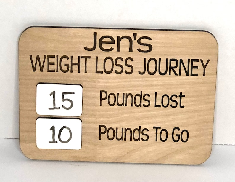 May include: A wooden sign with the text "Jen's Weight Loss Journey" and two white squares with the numbers "15" and "10" written in black marker. The text "Pounds Lost" and "Pounds To Go" are written below the squares.