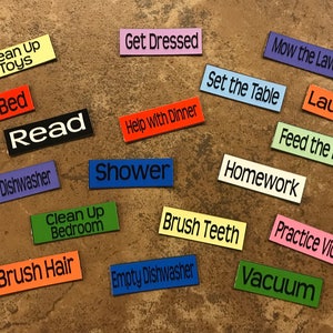 May include: A collection of colorful rectangular labels with various household chores written on them, such as "Clean Up Toys", "Make Bed", "Read", "Get Dressed", "Help with Dinner", "Shower", "Mow the Lawn", "Set the Table", "Laundry", "Feed the Pet", "Homework", "Clean Up Bedroom", "Brush Teeth", "Empty Dishwasher", "Practice Violin", "Vacuum", and "Brush Hair".